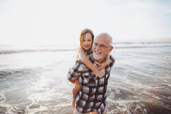 Fun, Energetic Grandpa Playing In Waves With Young Grandchild - Girl - On Beach At Sunset - Shoulder Ride